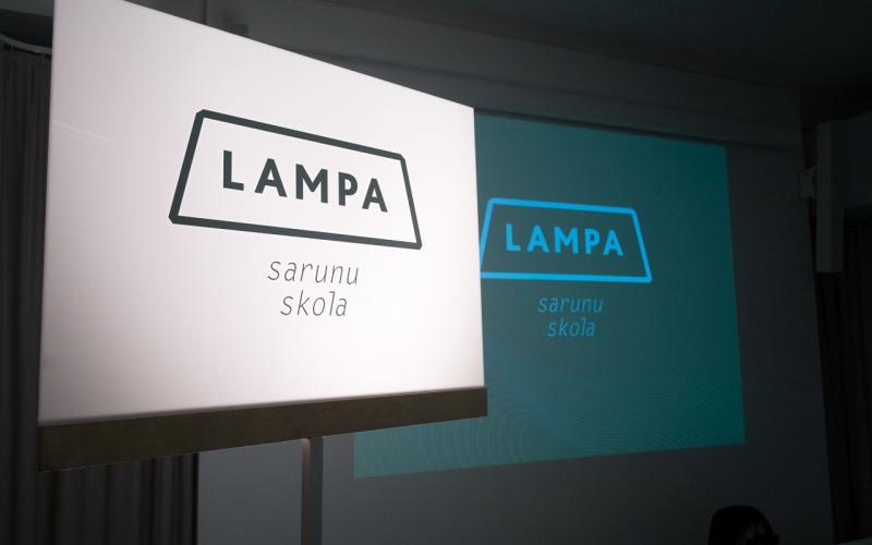 Izgaismota zīme ar tekstu "LAMPA sarunu skola" telpā ar projekciju, kurā redzams tas pats teksts. Atmosfēra ir izglītojoša un profesionāla.