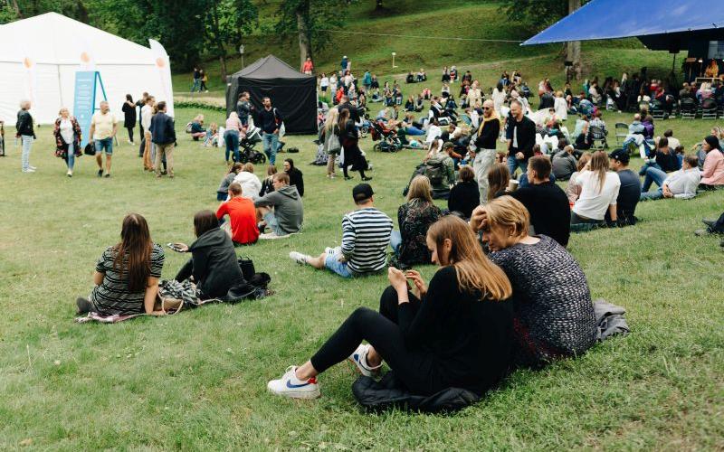 Brīvdabas festivāla aina ar cilvēkiem, kas sēž parka zālē, vērsti pret skatuvi. Redzamas teltis, valda relaksēta, kopīga atmosfēra zem mākoņainām debesīm.