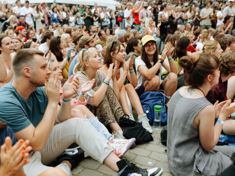 Dažādu cilvēku pūlis sēž ārā uz ietves, aplaudē un smaida, radot svētku un priecīgu atmosfēru festivālā.