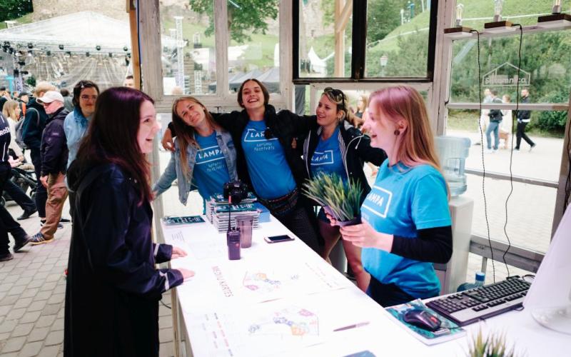 Četri smaidoši Sarunu festivāls LAMPA brīvprātīgie zilos kreklos stāv aiz informācijas stenda, sirsnīgi sarunājoties ar apmeklētāju. Atmosfēra ir rosīga un atrodas brīvā dabā.