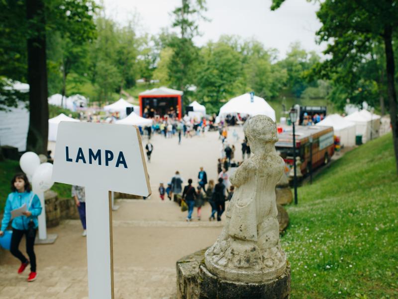 Dzīvīga parka aina ar norādes zīmi "LAMPA", akmens statuju un pūļiem, kas sapulcējušies pie baltām teltīm un skatuves.