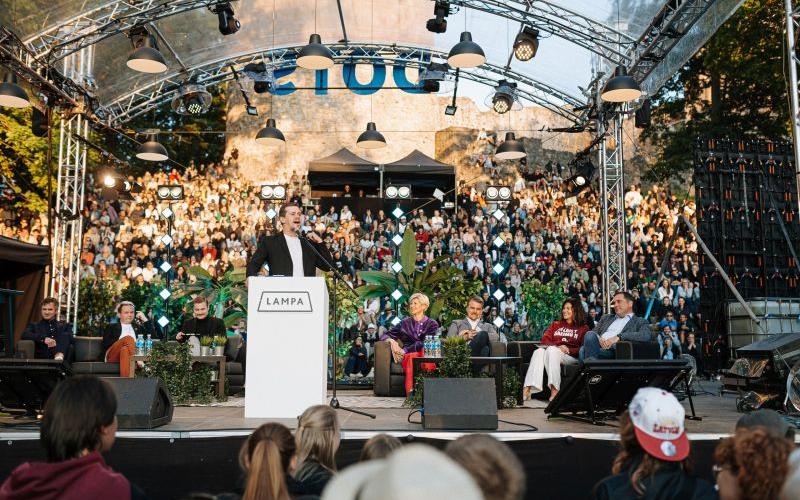 Komiķis Rūdolfs Kugrēns no tribīnes ar uzrakstu "LAMPA" uzrunā sēdošu paneli un lielu auditoriju ārā. Atmosfēra ir dzīvīga un saistoša.