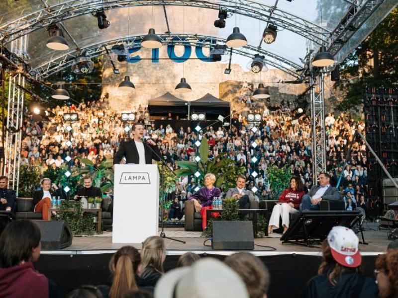 Komiķis Rūdolfs Kugrēns no tribīnes ar uzrakstu "LAMPA" uzrunā sēdošu paneli un lielu auditoriju ārā. Atmosfēra ir dzīvīga un saistoša.