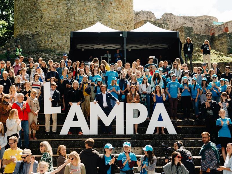 Uz pakāpieniem pulcējās dažādu cilvēku pūlis, pozējot ar lieliem burtiem "LAMPA". Gaiša vasaras diena, dzīvespriecīga atmosfēra, fonā pilsdrupas.