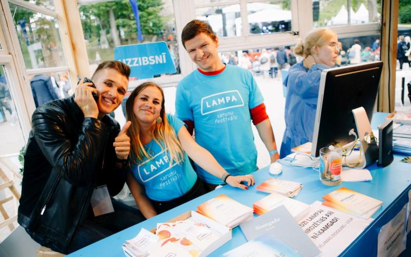 Trīs smaidoši cilvēki pie festivāla stenda ģērbušies zilos "LAMPA" kreklos, uz galda atrodas bukleti. Viens cilvēks tur rokās telefonu, rādot īkšķi uz augšu. Noskaņojums ir dzīvīgs un viesmīlīgs.
