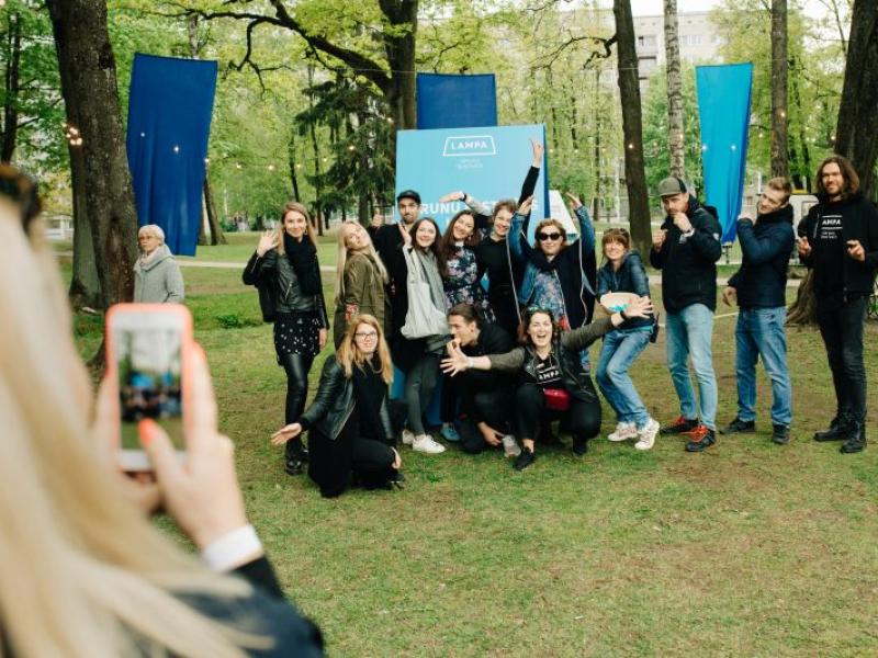 Priecīgu cilvēku grupa pozē fotogrāfijai zaļā parkā, daži paceļ rokas. Priekšplānā esoša persona iemūžina mirkli ar tālruni. Fonā redzami zili baneri un koki, radot svētku atmosfēru.