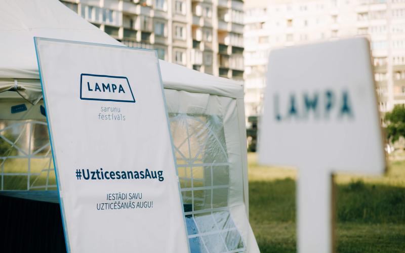 "Festivāla telts ar 'LAMPA' baneri un mirkļbirku #UzticesanasAug pilsētas parka vidē. Dzīvokļu ēkas izplūdušas fonā, saulaina atmosfēra."