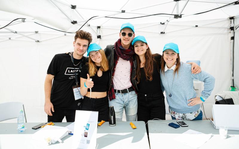 Piecu smaidīgu cilvēku grupa, kas stāv kopā zem baltas telts, ģērbušies ikdienas apģērbā un zilās cepurēs, izstarojot dzīvespriecīgu un draudzīgu atmosfēru.