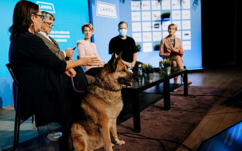 Paneļdiskusija ar pieciem sēdošiem cilvēkiem, vienam maskā, uz skatuves ar logotipu fonu. Priekšā sēž vācu aitu suns.