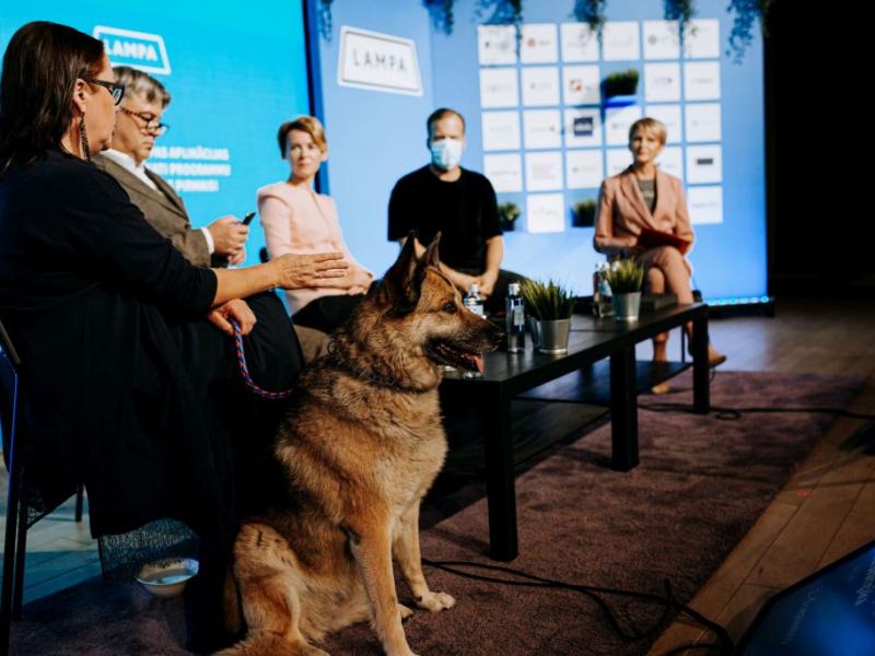 Paneļdiskusija ar pieciem sēdošiem cilvēkiem, vienam maskā, uz skatuves ar logotipu fonu. Priekšā sēž vācu aitu suns.