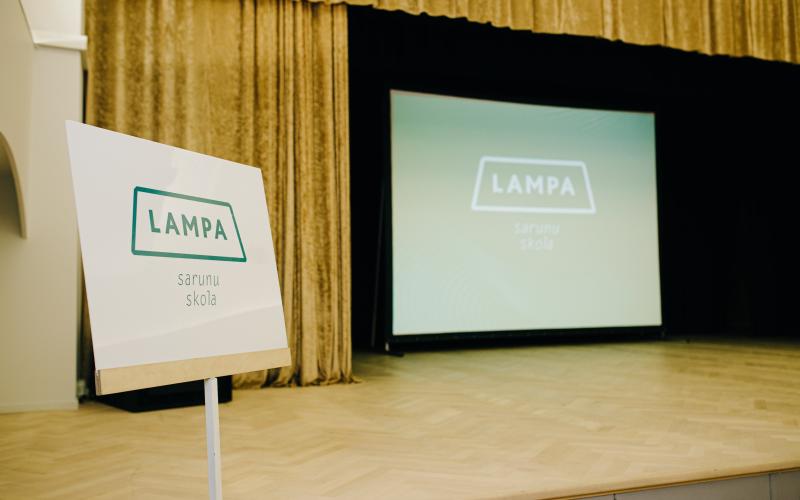 Blakus skatuvei, uz kuras ir liels ekrāns ar to pašu tekstu, stāv zīme ar uzrakstu "LAMPA sarunu skola". Iestāde atrodas silti apgaismotā auditorijā, kas rada gaidas.
