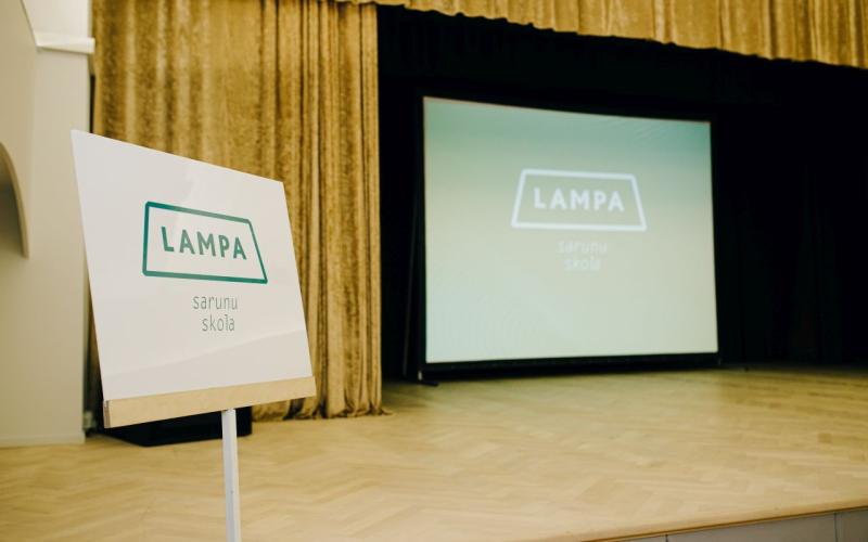Blakus skatuvei, uz kuras ir liels ekrāns ar to pašu tekstu, stāv zīme ar uzrakstu "LAMPA sarunu skola". Iestāde atrodas silti apgaismotā auditorijā, kas rada gaidas.