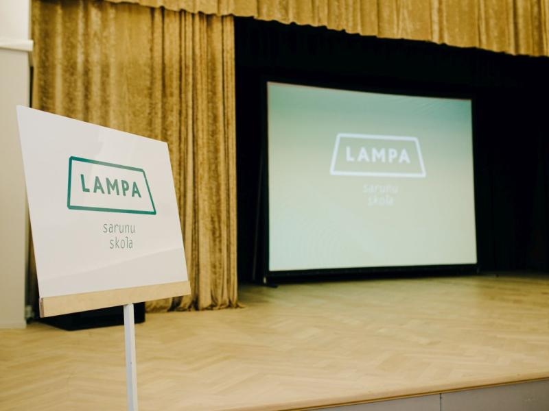 Blakus skatuvei, uz kuras ir liels ekrāns ar to pašu tekstu, stāv zīme ar uzrakstu "LAMPA sarunu skola". Iestāde atrodas silti apgaismotā auditorijā, kas rada gaidas.