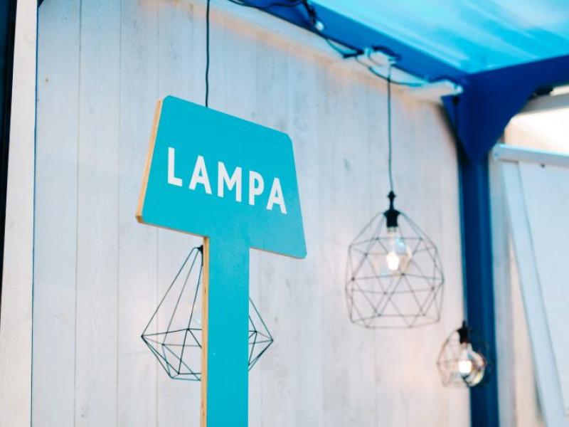 Zila zīme ar baltu uzrakstu "LAMPA" uz gaišas koka sienas. Netālu atrodas ģeometriskas piekaramās gaismas, kas rada modernu un gaisīgu atmosfēru.