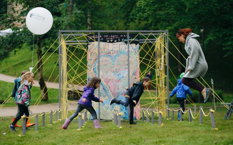 Bērnu grupa un pieaugušais parkā priecīgi skrien un lec krāsainas sirds formas sienas gleznojuma priekšā. Kreisajā pusē redzams balons, un tos ieskauj zaļi koki.