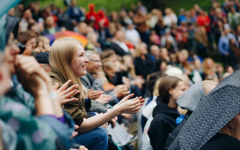 Cilvēku pūlis sēž ārā, aplaudē un smaida. Fokusā ir sieviete ar blondiem matiem, ko ieskauj krāsaini lietussargi un izplūdušas figūras, radot dzīvīgu, dzīvespriecīgu atmosfēru.