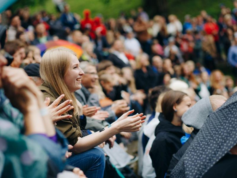 Cilvēku pūlis sēž ārā, aplaudē un smaida. Fokusā ir sieviete ar blondiem matiem, ko ieskauj krāsaini lietussargi un izplūdušas figūras, radot dzīvīgu, dzīvespriecīgu atmosfēru.