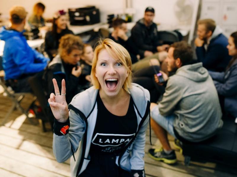 Priekšplānā priecīga sieviete rāda miera zīmi, plati smaidot. Fonā cilvēku grupa mierīgi sēž, sarunājas un ir atslābinājusies.