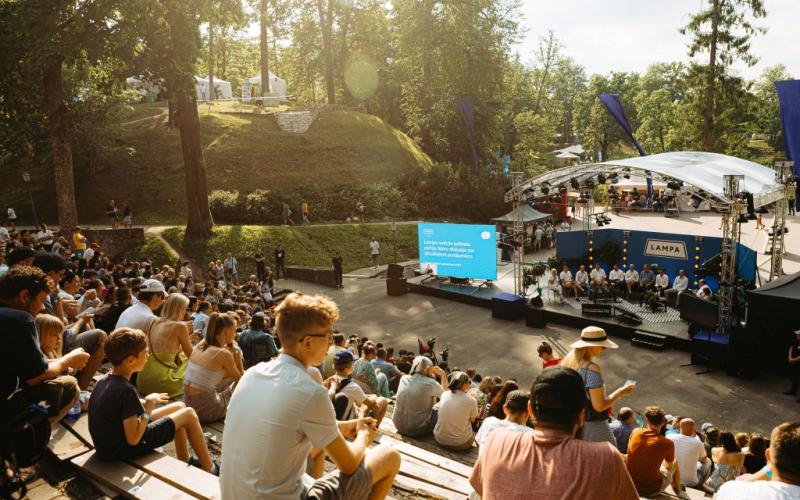 Āra pasākums parkā ar lielu auditoriju, kas sēž uz pakāpieniem ar skatuves skatuvi. Saulaina diena, koki fonā un liels ekrāns ar tekstu.