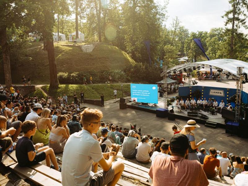 Āra pasākums parkā ar lielu auditoriju, kas sēž uz pakāpieniem ar skatuves skatuvi. Saulaina diena, koki fonā un liels ekrāns ar tekstu.