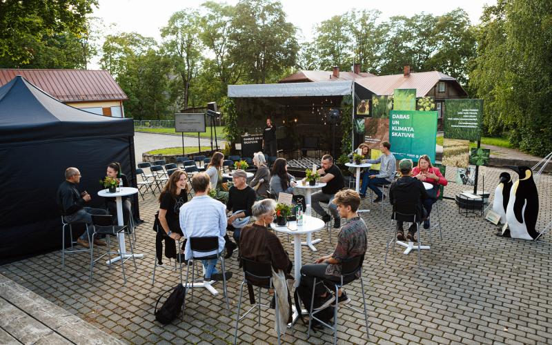 Neformāla pulcēšanās brīvā dabā, kur cilvēki sēž pie nelieliem galdiņiem bruģētā laukumā. Fonā uz skatuves uzstājas runātājs. Apstādījumi un relaksēta atmosfēra.