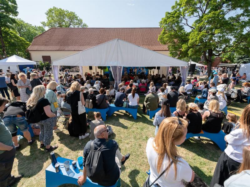 Brīvdabas festivāla aina ar pūli, kas sēž uz ziliem soliem un stāv zem baltas nojumes. Koki un zilas debesis rada dzīvīgu atmosfēru.