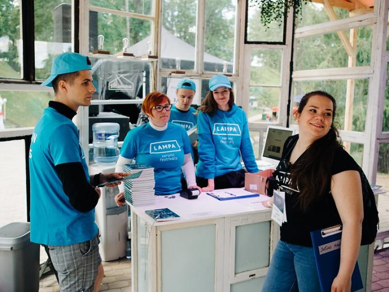 Festivāla brīvprātīgo grupa vienādos zilos formas tērpos stāv pie informācijas stenda. Vide ir gaiša un mājīga.