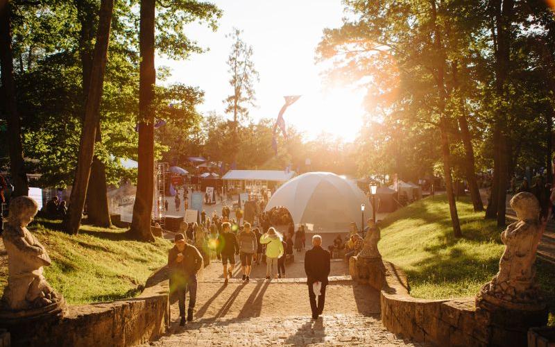 Saulains parka skats ar cilvēkiem, kas iet pa akmens taku kupola formas telts virzienā. Taku ieskauj augsti koki, radot siltu un aicinošu atmosfēru.