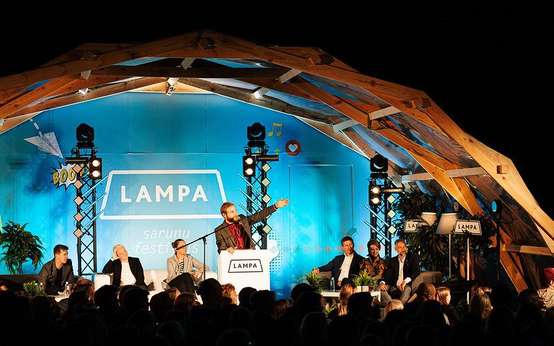 Spilgts runātāju panelis festivālā zem koka arkas. Cilvēks pie tribīnes dzīvīgi žestikulē. Auditorija ir iesaistīta. Aiz muguras treknrakstā uzraksts "LAMPA".