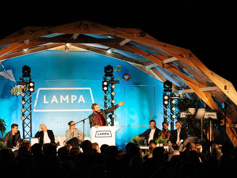 Spilgts runātāju panelis festivālā zem koka arkas. Cilvēks pie tribīnes dzīvīgi žestikulē. Auditorija ir iesaistīta. Aiz muguras treknrakstā uzraksts "LAMPA".