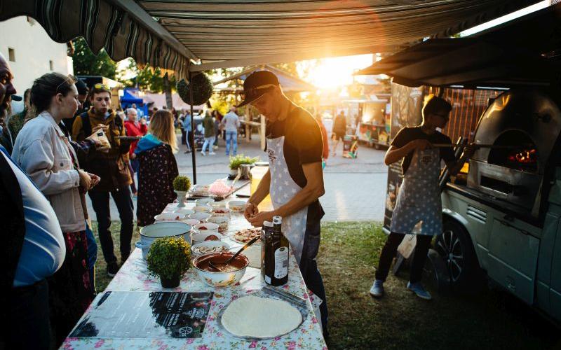 Āra ēdienu stends saulrietā ar vīrieti, kurš gatavo picas mīklu. Sastāvdaļas ir glīti izkārtotas uz ziedu raksta galdauta. Cilvēki nepacietīgi vēro.