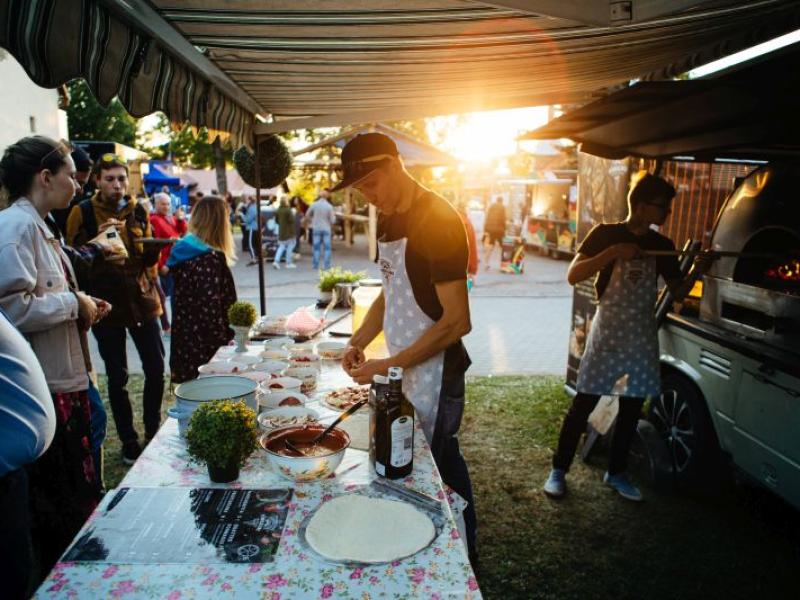 Āra ēdienu stends saulrietā ar vīrieti, kurš gatavo picas mīklu. Sastāvdaļas ir glīti izkārtotas uz ziedu raksta galdauta. Cilvēki nepacietīgi vēro.