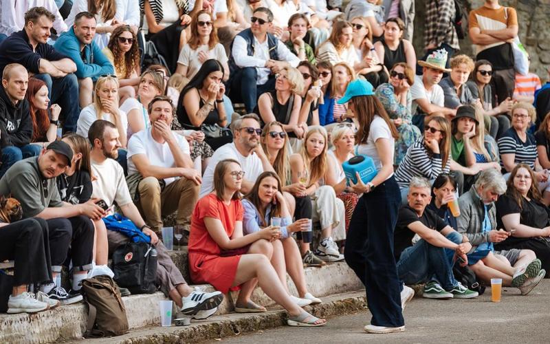 Cilvēku pūlis sēž uz āra kāpnēm un uzmanīgi vēro pasākumu. Sieviete ar zilu cepuri entuziastiski komunicē, radot aizraujošu un dzīvīgu atmosfēru.