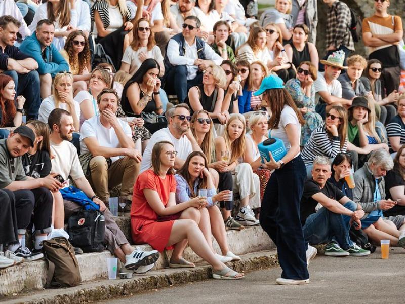 Cilvēku pūlis sēž uz āra kāpnēm un uzmanīgi vēro pasākumu. Sieviete ar zilu cepuri entuziastiski komunicē, radot aizraujošu un dzīvīgu atmosfēru.