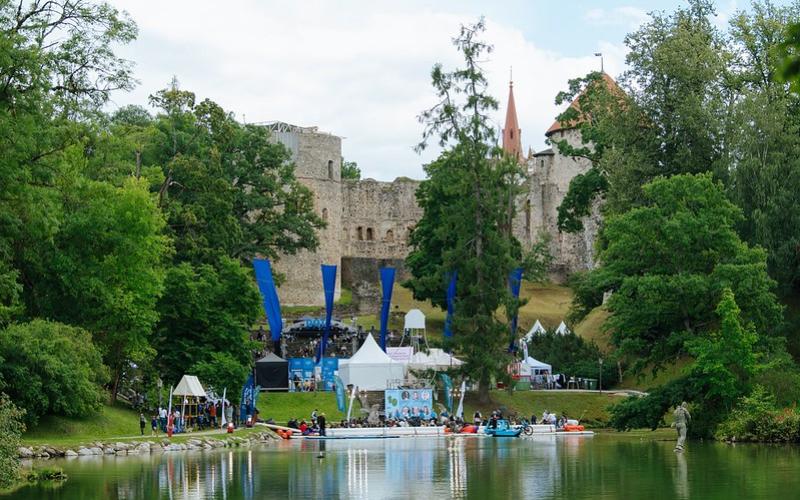 Cēsu pils pilsdrupas, ko ieskauj sulīgi zaļi koki zem mākoņainas debess. Cilvēki pulcējas pie ezera, teltis un baneri norāda uz pasākumu brīvā dabā.