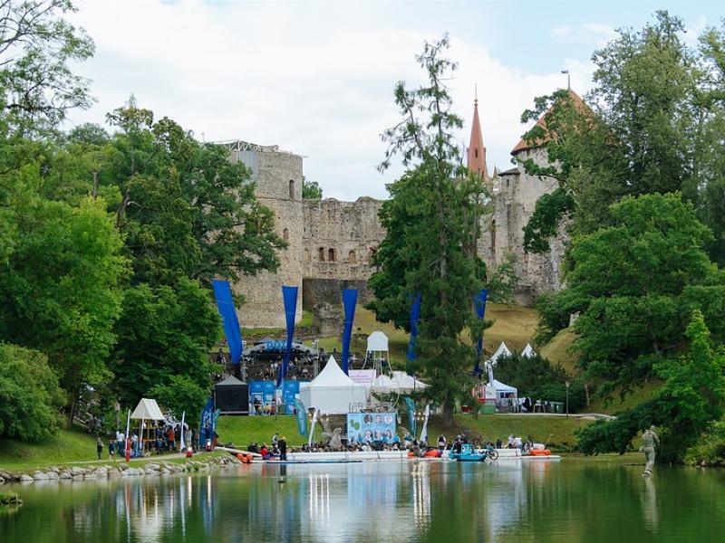 Cēsu pils pilsdrupas, ko ieskauj sulīgi zaļi koki zem mākoņainas debess. Cilvēki pulcējas pie ezera, teltis un baneri norāda uz pasākumu brīvā dabā.