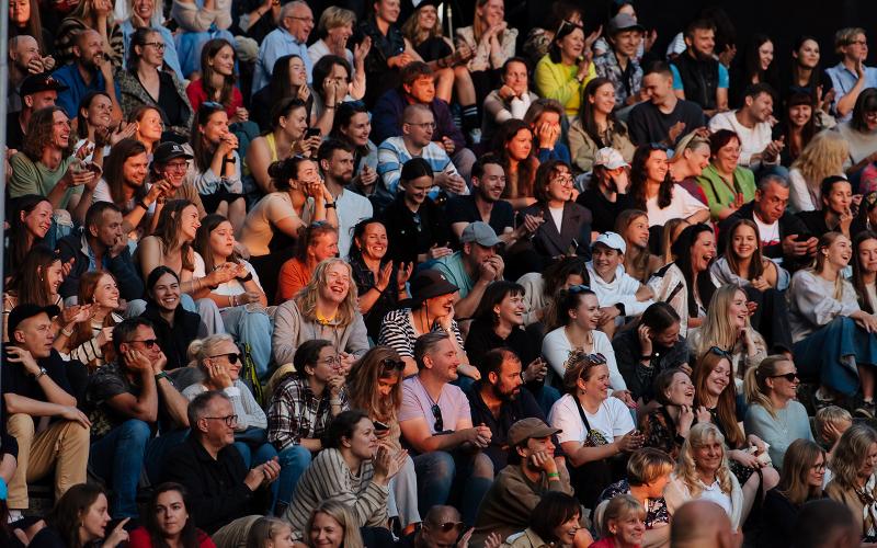 Liels, daudzveidīgs cilvēku pūlis, kas cieši sēž kopā, smaida un aplaudē. Noskaņojums ir priecīgs un dzīvīgs.