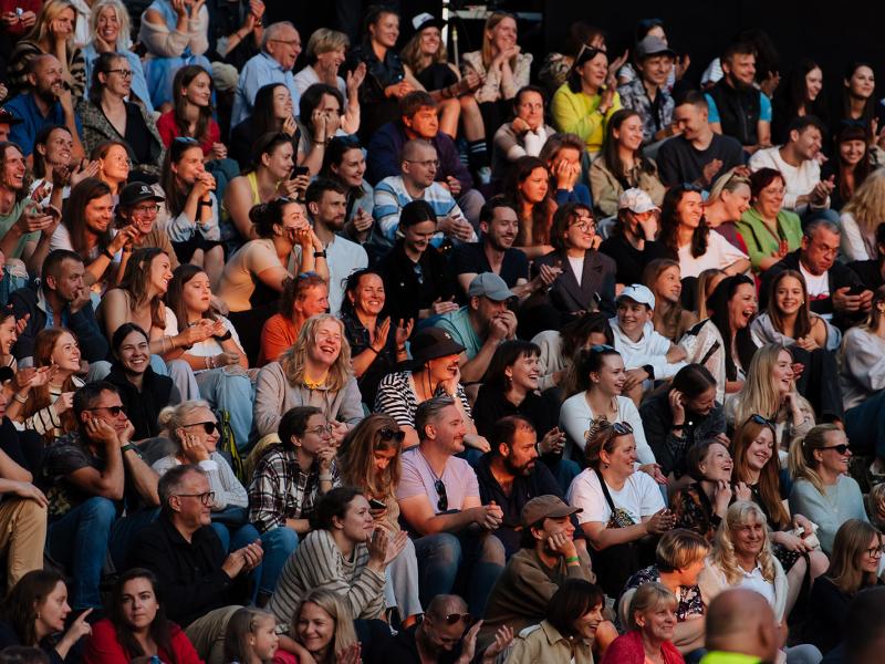 Liels, daudzveidīgs cilvēku pūlis, kas cieši sēž kopā, smaida un aplaudē. Noskaņojums ir priecīgs un dzīvīgs.