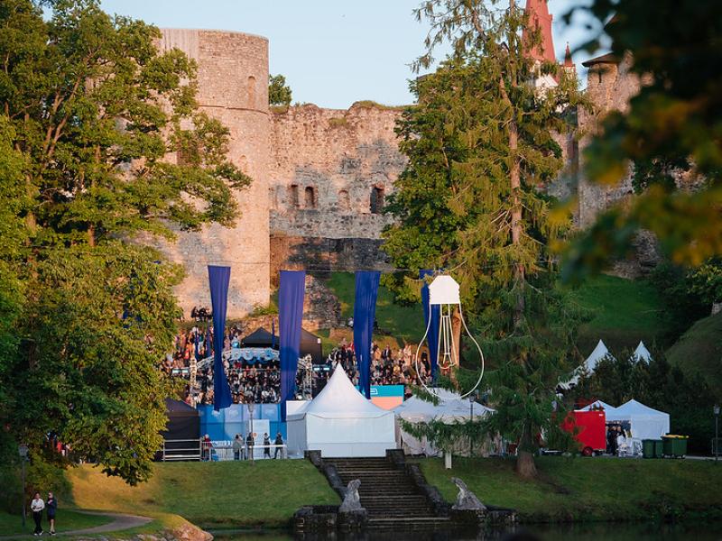 Cēsu pils pilsdrupām priekšā notiek brīvdabas festivāls ar ziliem karogiem, baltām teltīm un dzīvīgu pūli, ko ieskauj sulīga zaļums.