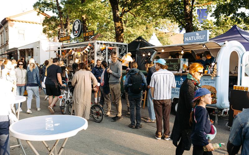 Festivāla ēdināšanas zonas aina, kurā cilvēki pulcējas ap ēdienu furgoniem zem lapu kokiem. Neformāla, dzīvīga atmosfēra. Galdiņi ir novietoti uz celiņa.