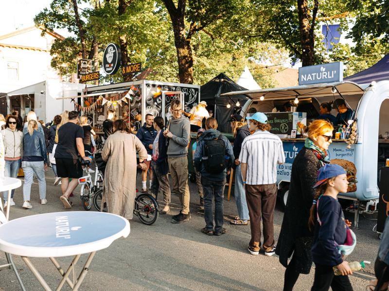 Festivāla ēdināšanas zonas aina, kurā cilvēki pulcējas ap ēdienu furgoniem zem lapu kokiem. Neformāla, dzīvīga atmosfēra. Galdiņi ir novietoti uz celiņa.