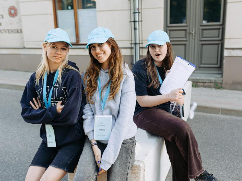Trīs jaunas sievietes - festivāla brīvprātīgās -zilās cepurēs pozē ikdienišķi. Viena tur rokās planšeti.
