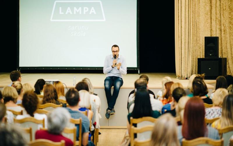 Silti apgaismotā telpā uz skatuves sēž runātājs ar mikrofonu rokās, uzrunājot auditoriju. Fonā redzamajā ekrānā ir redzams uzraksts "LAMPA".