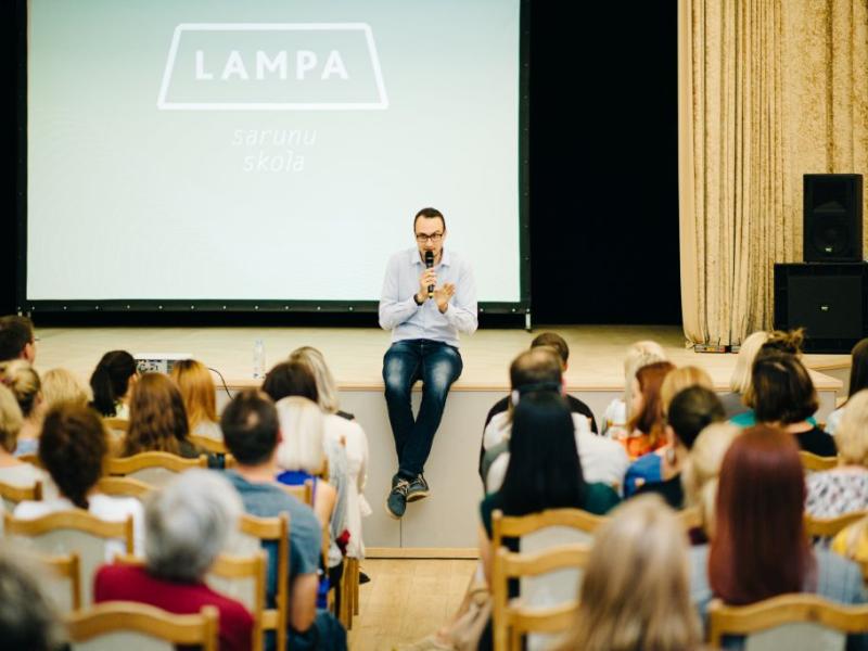 Silti apgaismotā telpā uz skatuves sēž runātājs ar mikrofonu rokās, uzrunājot auditoriju. Fonā redzamajā ekrānā ir redzams uzraksts "LAMPA".
