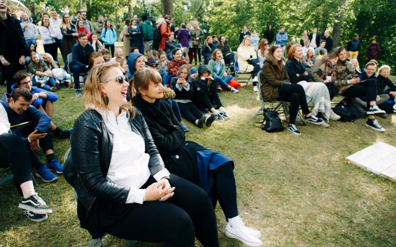 Dzīvīga publika brīvā dabā sēž zālē, smejas un bauda pasākumu. Koki un zaļumi rada mierīgu fonu. Pūlis ir atslābināts un iesaistīts.