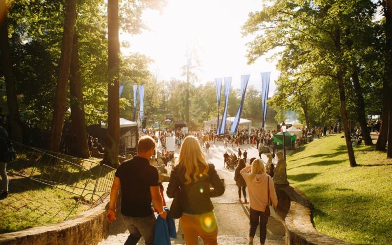 Saulains parka skats ar cilvēkiem, kas iet pa akmens taku. Taku ieskauj augsti koki, radot siltu un aicinošu atmosfēru.