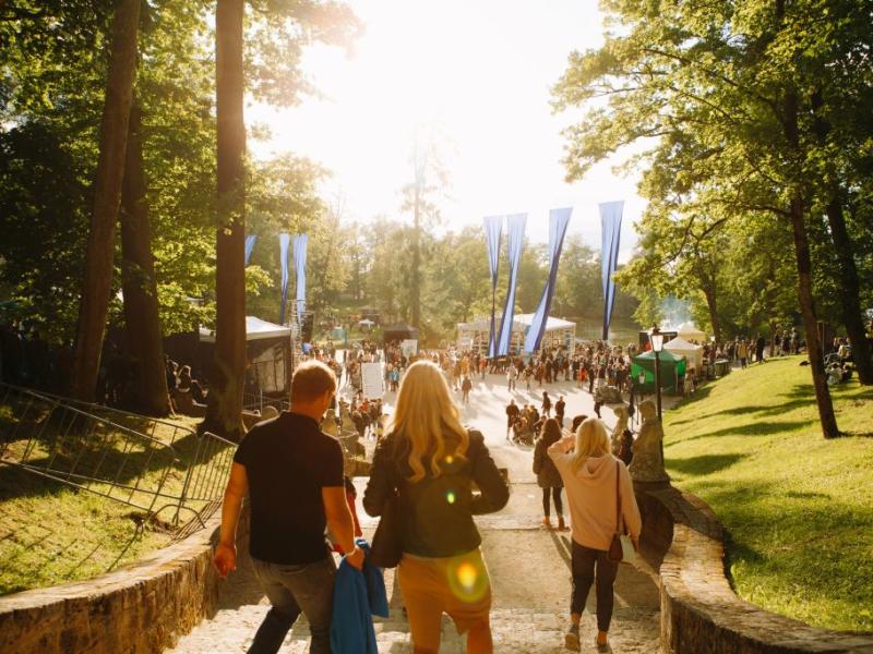 Saulains parka skats ar cilvēkiem, kas iet pa akmens taku. Taku ieskauj augsti koki, radot siltu un aicinošu atmosfēru.