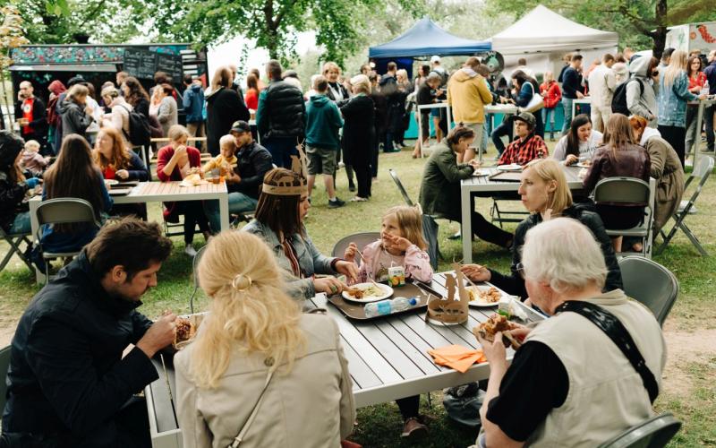 Festivāls, kurā visu vecumu cilvēki sēž pie galdiem un bauda maltītes zem kokiem. Fonā ir ēdienu furgoni un teltis, kas rada dzīvīgu atmosfēru.