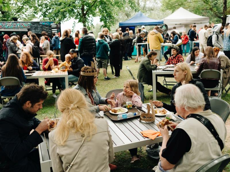 Festivāls, kurā visu vecumu cilvēki sēž pie galdiem un bauda maltītes zem kokiem. Fonā ir ēdienu furgoni un teltis, kas rada dzīvīgu atmosfēru.