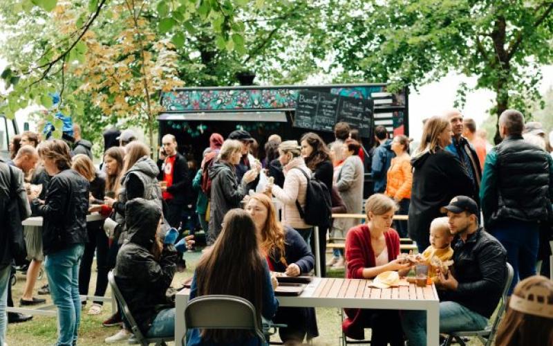 Festivāls, kurā visu vecumu cilvēki sēž pie galdiem un bauda maltītes zem kokiem. Fonā ir ēdienu furgoni un teltis, kas rada dzīvīgu atmosfēru.